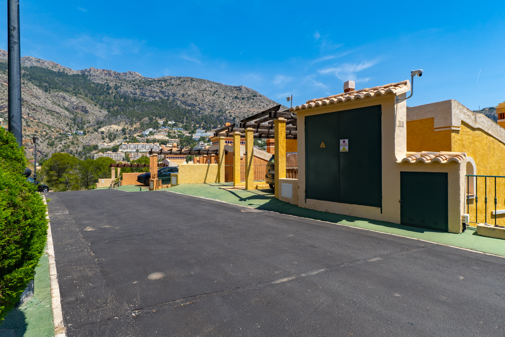 Calle de acceso residencial con edificio de servicios amarillo, cámara de seguridad y pérgola, enmarcada por una sierra verde con viviendas bajo un cielo azul despejado.