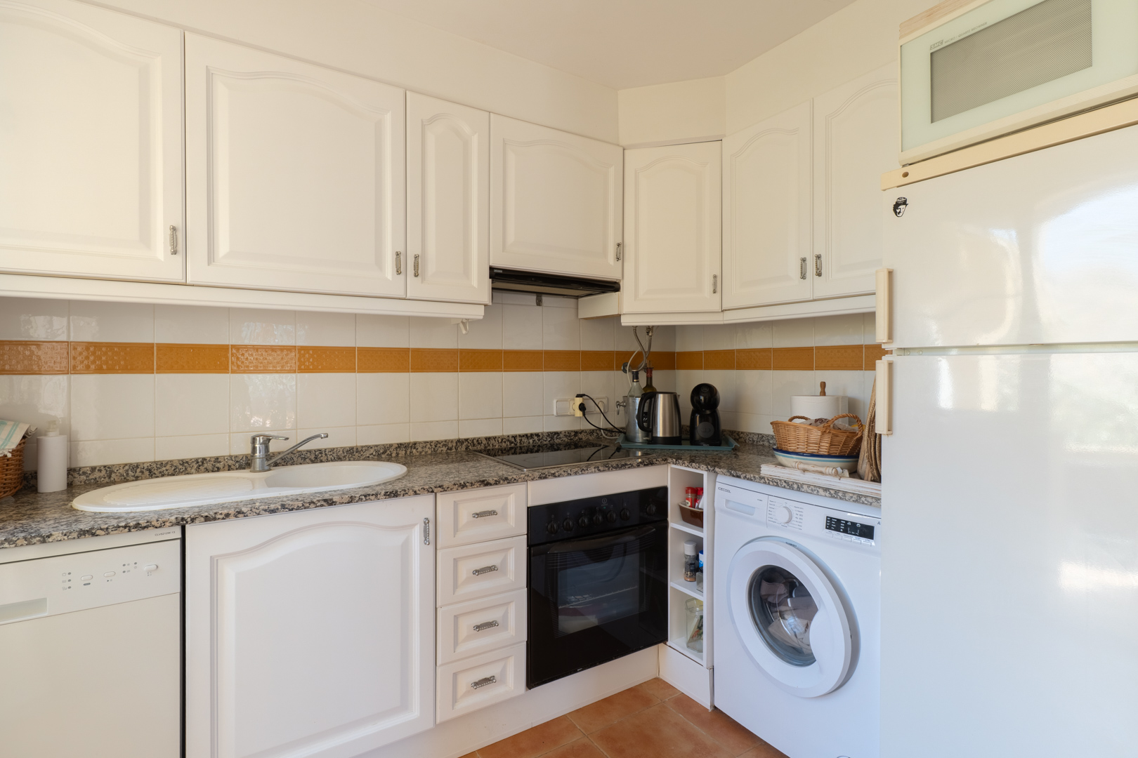 Cocina funcional con armarios blancos de paneles, encimera de granito, electrodomésticos integrados y azulejos con franja decorativa naranja.