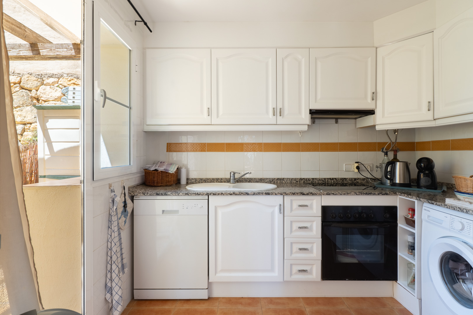 Cocina funcional con armarios blancos, encimera de granito y electrodomésticos integrados. Ventana abierta a un patio exterior con paredes de piedra y pérgola de madera.