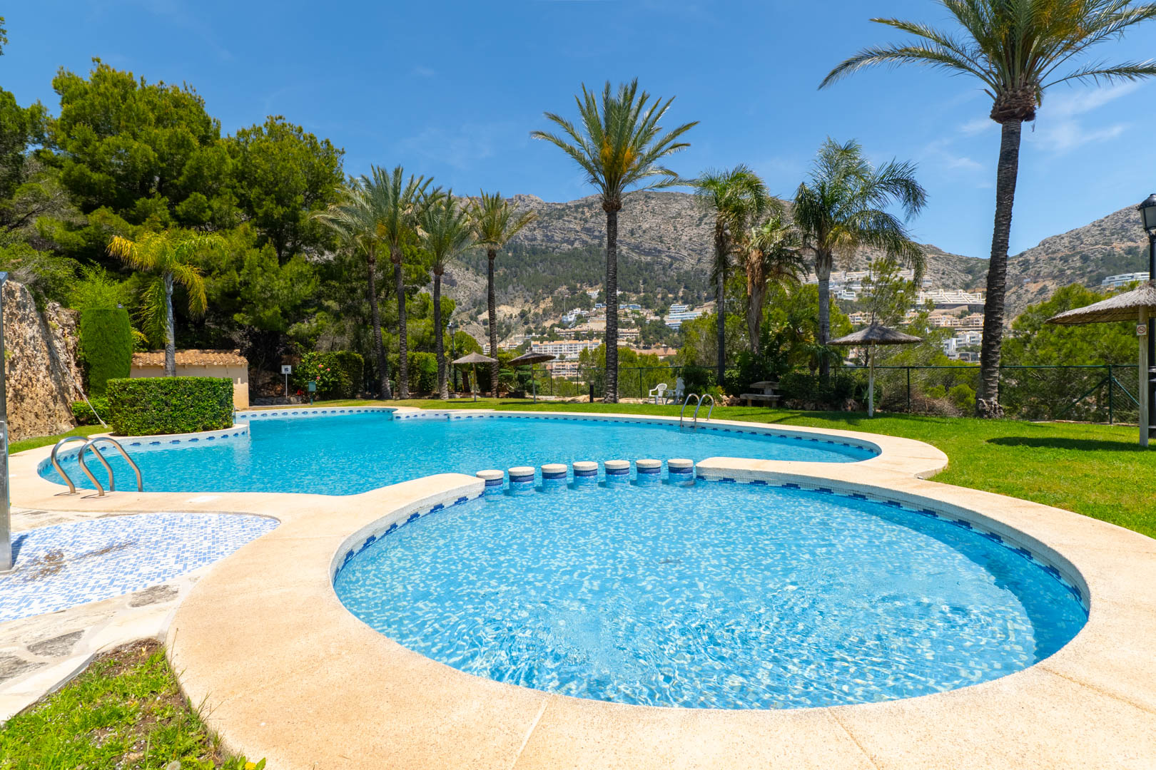 Complejo de piscinas de varios niveles con área infantil, rodeado de palmeras y césped. Ofrece vistas panorámicas a las montañas y la vegetación local bajo un cielo azul despejado.