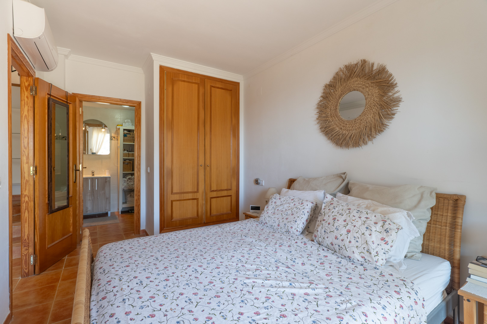 Dormitorio con cama de cabecero de mimbre, armario empotrado de madera clara y suelo de terracota. Se aprecia un espejo de fibras naturales y acceso a un baño.