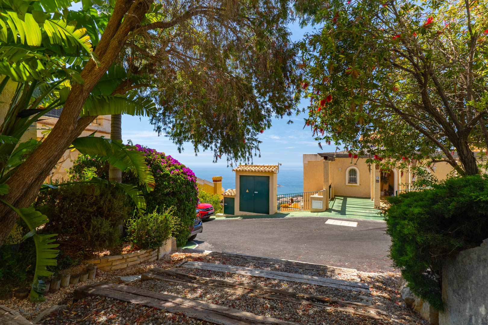 Entrada a la propiedad con camino pavimentado y sendero de grava con traviesas de madera, enmarcado por exuberante vegetación mediterránea y una vista parcial al mar.