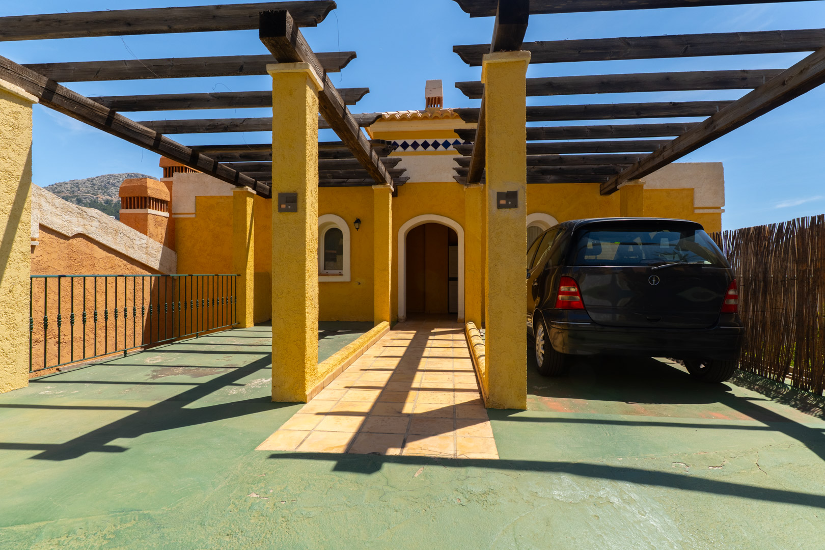 Entrada exterior con fachada de estuco amarillo y pérgola de madera sobre zona de aparcamiento verde. Incluye puerta arqueada, camino de baldosas y coche negro estacionado bajo la sombra.