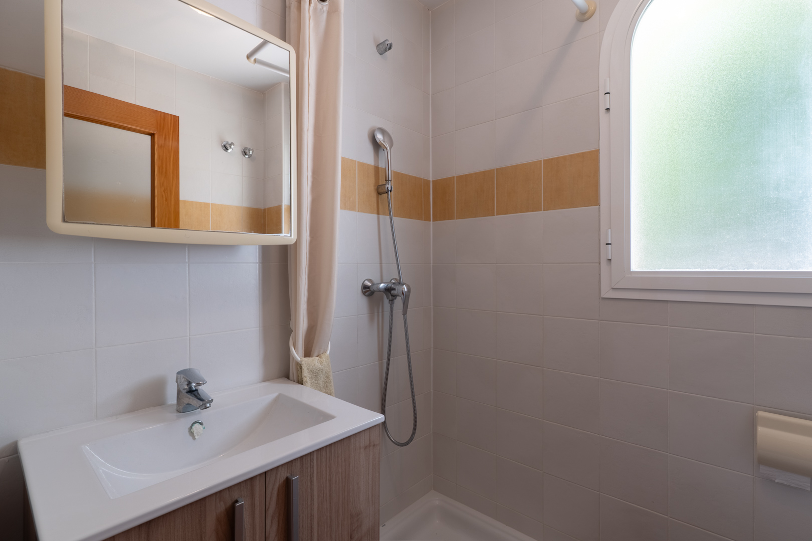Este baño funcional presenta un mueble de lavabo de madera clara, ducha con cortina y una ventana arqueada de cristal esmerilado, complementado con azulejos blancos y una banda decorativa ocre.