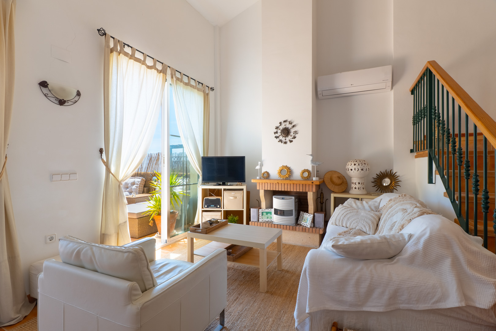 Sala de estar luminosa con techos altos, acceso a terraza, chimenea de ladrillo decorativa y escalera de madera con barandilla verde.