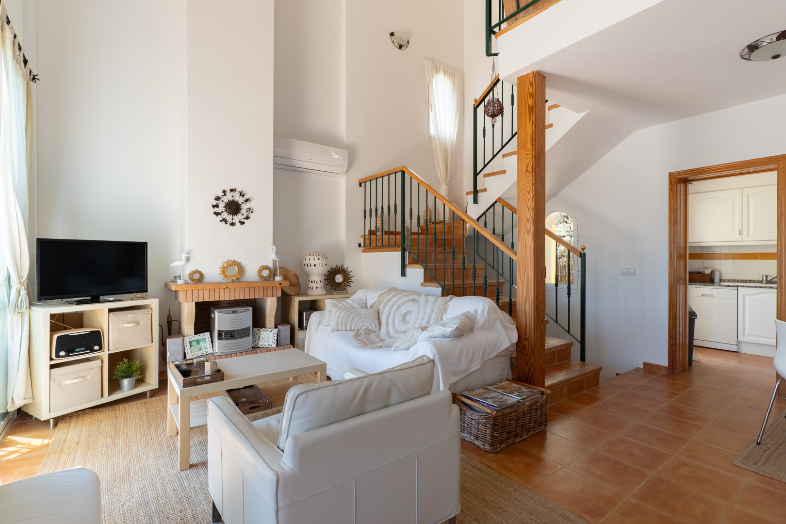 Salón acogedor con chimenea de ladrillo y madera, sofá blanco y escalera de madera con barandilla verde, conectando con la cocina.