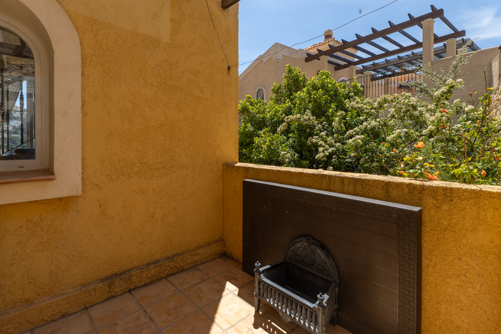 Terraza exterior con paredes de estuco ocre y suelo de baldosas. Presenta una ventana arqueada, un panel decorativo oscuro y vistas a jardines con flores y una pérgola.