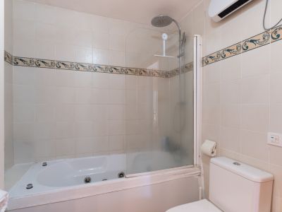 Baño completo con bañera de hidromasaje en esquina y ducha. Paredes alicatadas en tono beige con cenefa decorativa. Incluye inodoro y estantería de almacenamiento.