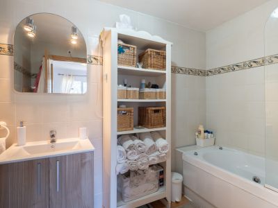 Baño moderno con lavabo sobre mueble de madera clara, espejo arqueado con focos y bañera. Incluye estantería blanca con cestas de mimbre y paredes alicatadas con cenefa decorativa.