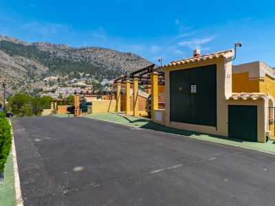 Calle de acceso residencial con edificio de servicios amarillo, cámara de seguridad y pérgola, enmarcada por una sierra verde con viviendas bajo un cielo azul despejado.