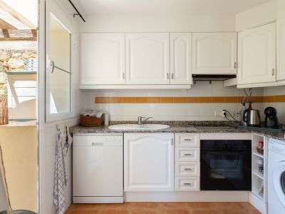 Cocina funcional con armarios blancos, encimera de granito y electrodomésticos integrados. Ventana abierta a un patio exterior con paredes de piedra y pérgola de madera.