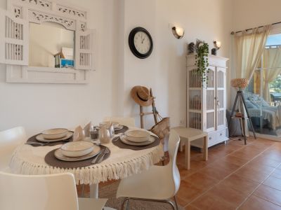 Comedor luminoso con mesa redonda, sillas blancas y un espejo decorativo de madera. El espacio se abre a una terraza exterior con vistas a las colinas, destacando suelos de terracota.