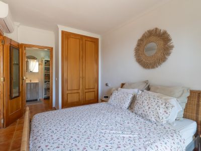 Dormitorio con cama de cabecero de mimbre, armario empotrado de madera clara y suelo de terracota. Se aprecia un espejo de fibras naturales y acceso a un baño.