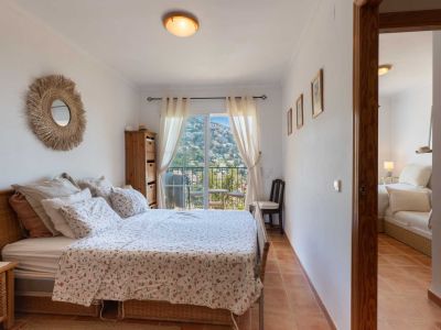 Dormitorio con cama de mimbre, suelo de terracota y acceso a balcón con vistas a la ladera. Incluye un espejo de rafia y muebles de madera clara.