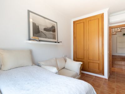 Dormitorio secundario con cama individual, sofá auxiliar, armario empotrado de madera y estantería con arte de playa. Suelos de terracota y acceso a pasillo con escaleras.