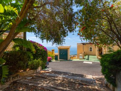 Entrada a la propiedad con camino pavimentado y sendero de grava con traviesas de madera, enmarcado por exuberante vegetación mediterránea y una vista parcial al mar.
