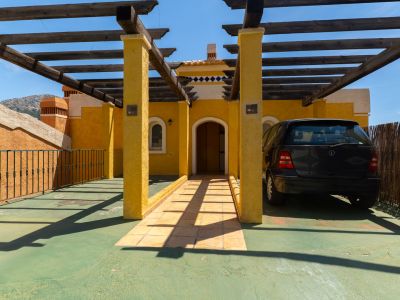 Entrada exterior con fachada de estuco amarillo y pérgola de madera sobre zona de aparcamiento verde. Incluye puerta arqueada, camino de baldosas y coche negro estacionado bajo la sombra.