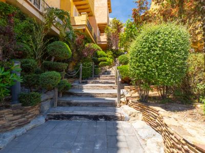 Escalera exterior de madera y piedra, flanqueada por exuberante vegetación mediterránea y muros de contención, que asciende hacia un edificio ocre con balcones.