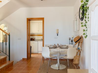 Luminoso comedor con mesa redonda y sillas modernas, suelo de baldosas de terracota y cocina compacta adyacente. Escalera de madera con ventana arqueada visible.