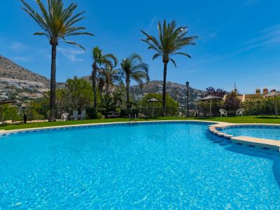 Piscina comunitaria de dos niveles con mosaicos azules, rodeada de césped y palmeras, ofreciendo un entorno relajante con vistas a las montañas de Altea Hills.