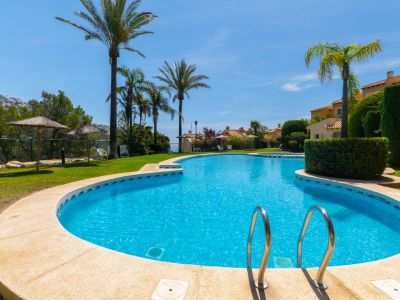 Piscina comunitaria de forma libre con borde de piedra y escaleras de acero, rodeada de césped, palmeras y sombrillas de paja, con vistas a colinas y viviendas.