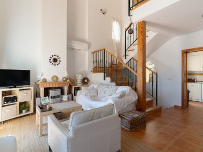 Salón acogedor con chimenea de ladrillo y madera, sofá blanco y escalera de madera con barandilla verde, conectando con la cocina.