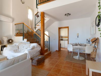 Salón-comedor diáfano con suelos de terracota y una escalera de madera con barandilla metálica. Incluye zona de estar con sofás blancos y comedor con mesa redonda.