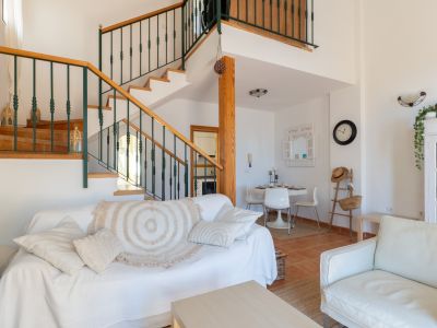 Salón luminoso con sofá blanco, mesa de centro de madera clara y una escalera de madera con barandilla verde oscuro. Incluye un área de comedor con mesa redonda.