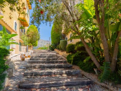Sendero exterior ascendente con escalones de traviesas de madera y grava, flanqueado por exuberante vegetación mediterránea. Un edificio amarillo con ventanas arqueadas se asoma a la izquierda.