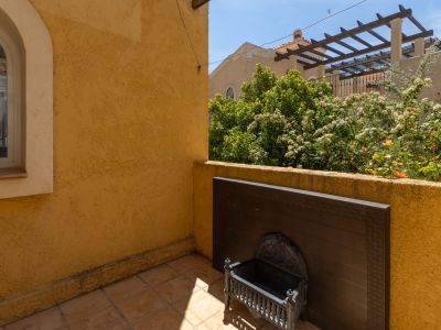 Terraza exterior con paredes de estuco ocre y suelo de baldosas. Presenta una ventana arqueada, un panel decorativo oscuro y vistas a jardines con flores y una pérgola.