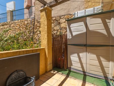 Terraza exterior con suelo de baldosas, armario de almacenamiento, muro de piedra con plantas florecidas y pérgola de madera, ofreciendo espacio funcional al aire libre.