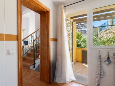 Vista desde un interior alicatado, con un marco de madera que da a una escalera de terracota y una puerta de cristal a una terraza exterior con muro de piedra y vegetación.