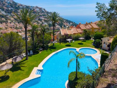 Zona de piscina comunitaria con gran piscina azul, césped verde, palmeras y sombrillas de paja. Ofrece vistas panorámicas al mar y a residencias adosadas en una ladera soleada.