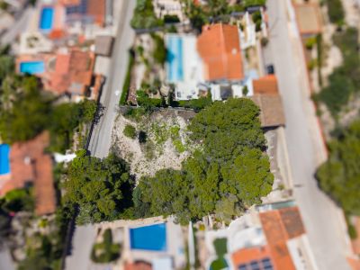 Vista aérea de una parcela sin desarrollar con árboles, rodeada de calles y propiedades residenciales con piscinas y tejados rojos.