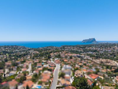 Vista aérea panorámica de la costa de Benissa, mostrando el mar Mediterráneo, el Peñón de Ifach y una zona residencial con vegetación exuberante bajo un cielo azul claro.
