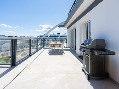 Amplia terraza con suelo de baldosas claras, barandillas de cristal y una moderna barbacoa. Incluye un comedor exterior y vistas a edificios y montañas.