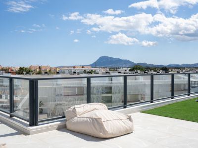 Amplia terraza en la azotea con suelo de gres y césped artificial, delimitada por una barandilla de cristal, ofreciendo vistas a la montaña y la ciudad.