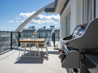 Amplia terraza en la azotea con una moderna barbacoa de gas y un juego de comedor exterior con mesa de madera, barandilla de cristal y suelo claro bajo cielo azul.
