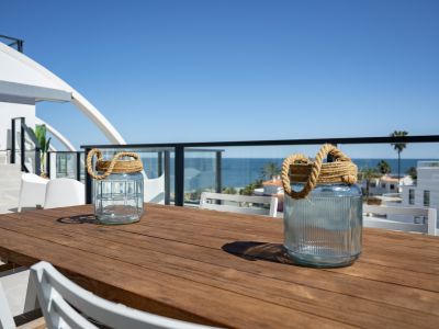 Amplia terraza exterior con mesa de madera y sillas blancas, ofreciendo vistas parciales al mar Mediterráneo y al entorno costero, bajo un cielo despejado.