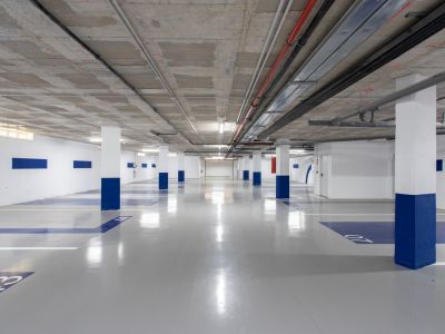 Amplio garaje subterráneo con techo de hormigón visto y conductos industriales. Pilares blancos con bases azules y suelo de epoxi gris con plazas numeradas. Iluminación fluorescente y cargador para vehículos eléctricos.