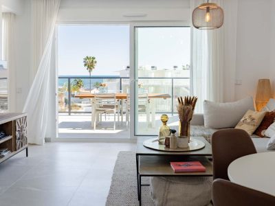 Amplio salón con suelo de baldosas claras y acceso directo a una terraza con vistas panorámicas al mar y una palmera. Incluye una lámpara colgante moderna y cortinas blancas translúcidas.