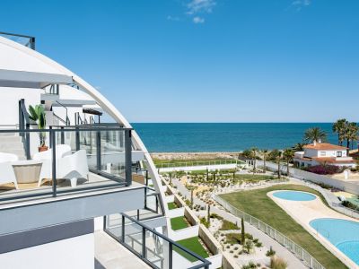 Balcón moderno con barandilla de cristal y mobiliario blanco, ofreciendo vistas panorámicas al mar Mediterráneo, piscinas comunitarias y jardines paisajísticos.