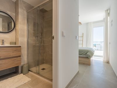 Baño moderno con azulejos efecto mármol, mueble de madera y ducha acristalada. Desde la entrada se aprecia el dormitorio con cama y vistas al mar.