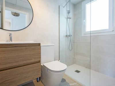 Baño moderno con mueble de lavabo flotante de madera clara, inodoro y ducha con mampara de cristal y ventana integrada, paredes de azulejos grises.