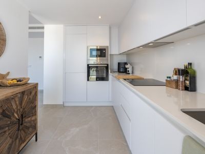 Cocina moderna con muebles blancos sin tiradores, electrodomésticos integrados de acero y encimeras claras. El suelo de gres porcelánico se complementa con una consola de madera rústica.