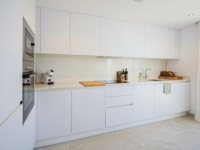 Cocina moderna y funcional con armarios blancos sin tiradores, encimera clara y electrodomésticos integrados. Incluye horno, microondas y placa de inducción, complementada con iluminación empotrada y suelo de gres claro.