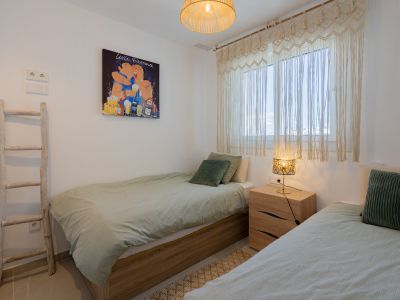 Dormitorio con dos camas individuales, marcos de madera clara y ropa de cama verde. Destacan las cortinas de macramé, una lámpara de mesa con pantalla decorada y una obra de arte pop en la pared.