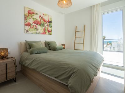 Dormitorio luminoso con cama de madera clara, ropa de cama verde salvia y un cuadro de flamencos. Gran ventanal con vistas parciales al mar.