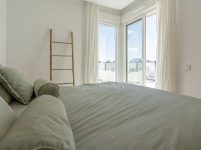 Dormitorio luminoso con cama vestida en tono salvia, acceso a terraza a través de amplios ventanales y escalera decorativa de madera.