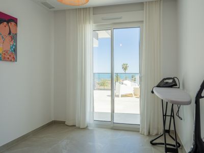 Estancia con suelo de gres y puerta corredera de cristal que da a una terraza con vistas al mar y palmeras. Se aprecian una tabla de planchar y una pintura colorida.